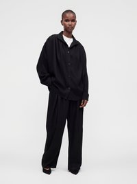 Personne portant une chemise noire ample boutonnée et un pantalon noir à larges jambes avec des chaussures pointues noires, debout devant un fond clair uni.