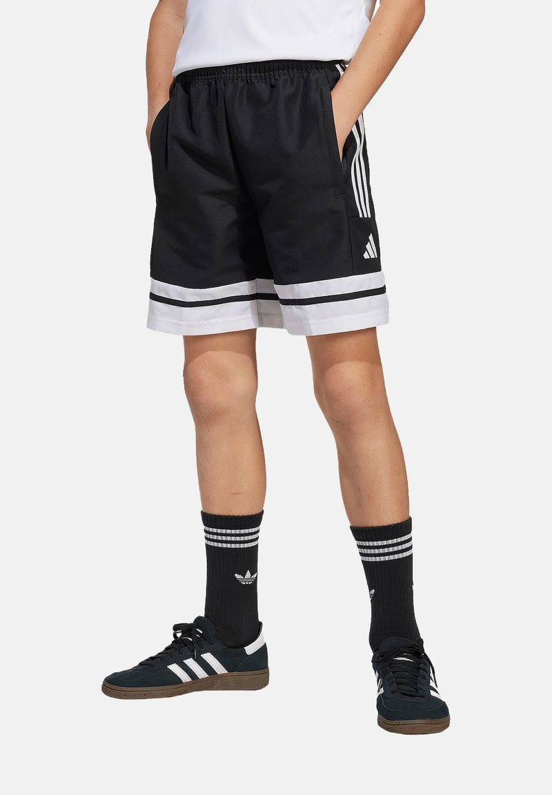 Zwarte sportshorts met witte strepen, twee zijzakken en een elastische tailleband. Gecombineerd met zwarte sokken en schoenen met witte accenten.