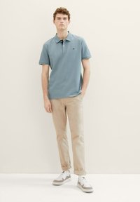 Polo bleu clair, en coton, à manches courtes, associé à un pantalon chino beige et des baskets grises et blanches, se tenant devant un arrière-plan neutre.