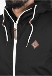 Veste à capuche noire dotée d'une fermeture éclair blanche, de cordons avec des accents en cuir et d'un petit patch logo marron sur la poitrine. Texture de tissu lisse.