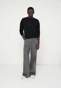 Sudadera negra con logotipo en relieve, combinada con pantalones grises de piernas anchas con cintura ajustable y rayas verticales. Zapatillas blancas completan el look.