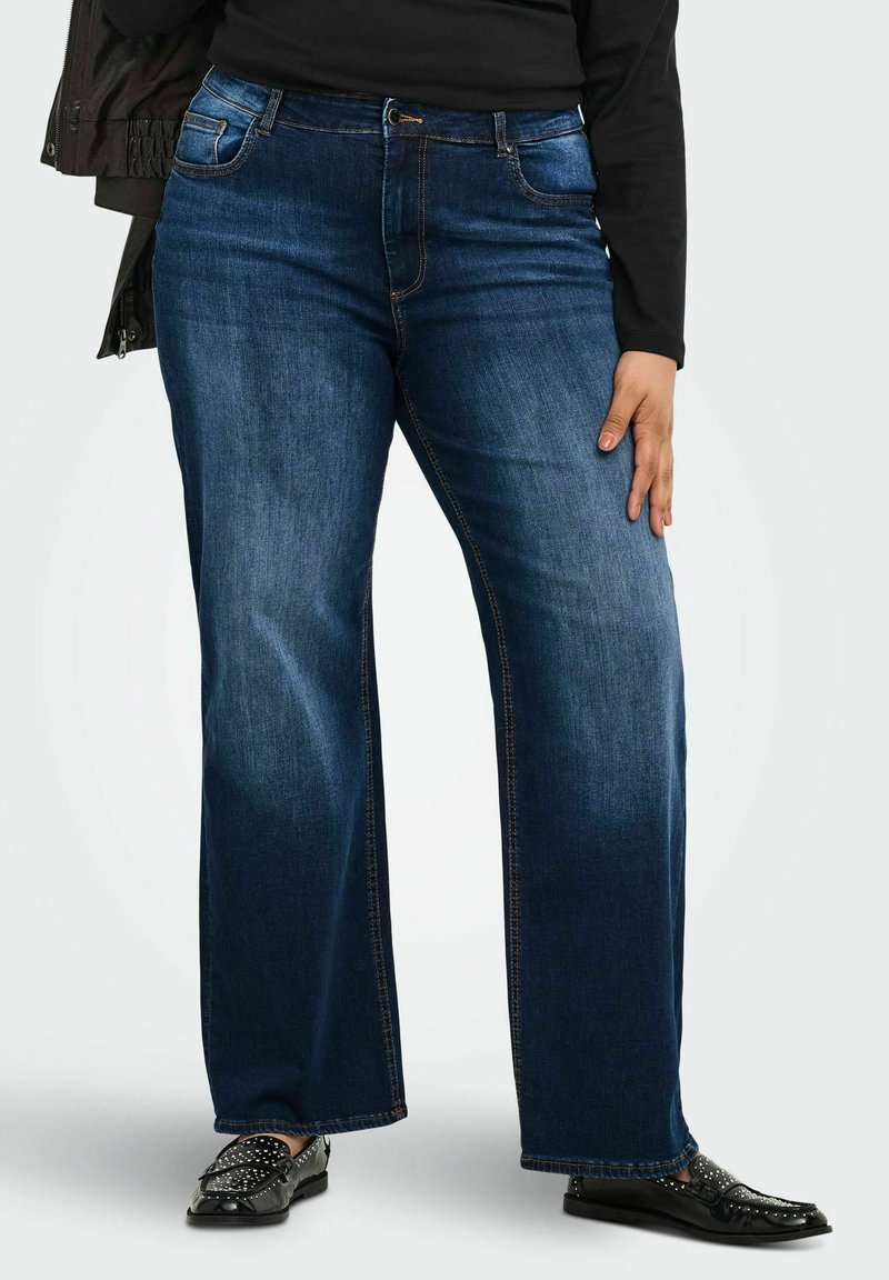 Άτομο που φορά σκούρα μπλε jeans με φαρδιά μπατζάκια, μαύρο μακρυμάνικο τοπ και μαύρα moccasins με τρουκς, στέκεται μπροστά σε απλό φόντο.