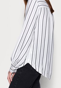 Blusa bianca con righe verticali grigie, maniche lunghe, tessuto morbido e un leggero orlo asimmetrico. Dettaglio di bottoni sui polsini.