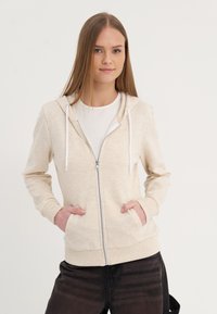 Felpa con zip beige con cappuccio, lacci bianchi, tasche frontali e polsini a coste, abbinata a pantaloni neri. Tessuto morbido.