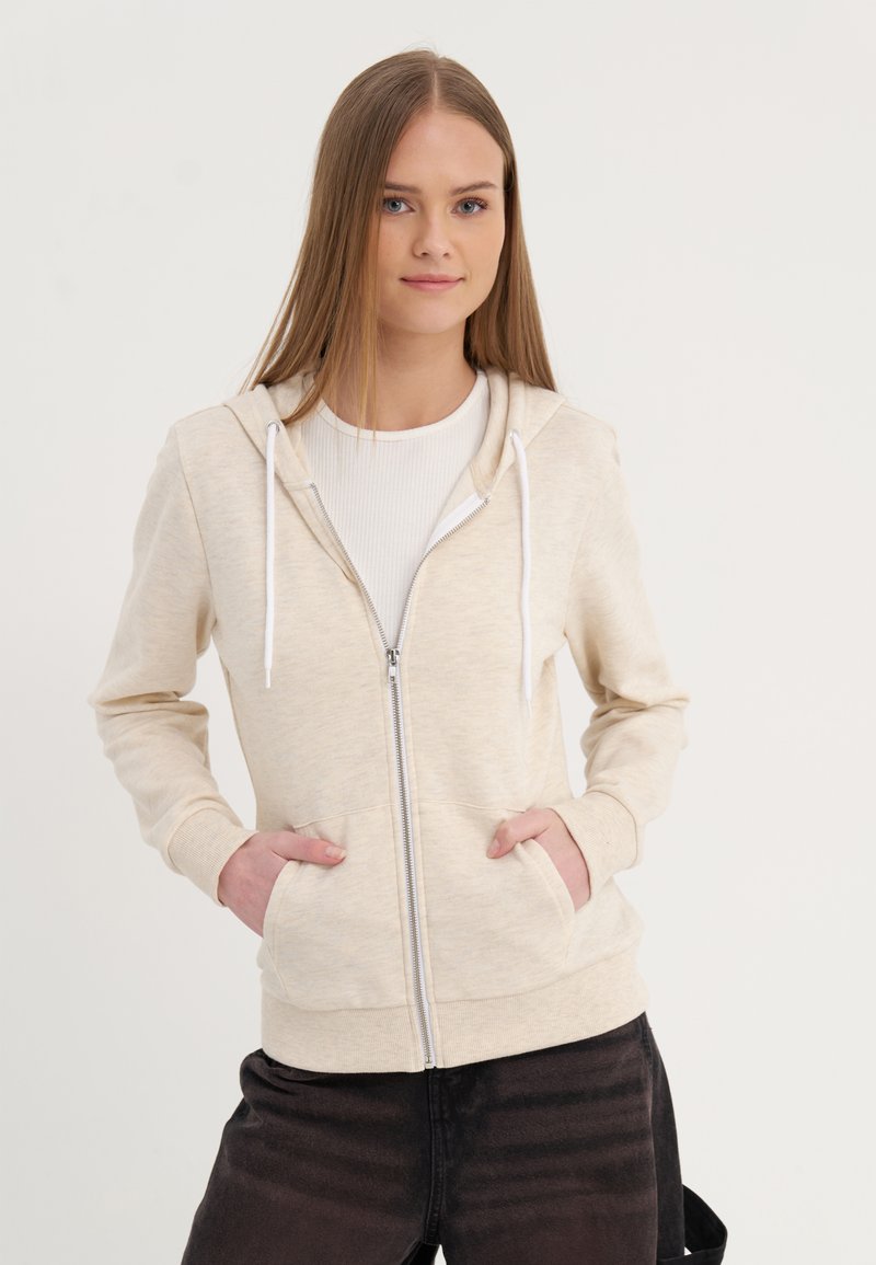 Sweatshirt bege com fecho éclair e capuz, cordões brancos, bolsos frontais e punhos canelados, combinado com calças pretas. Textura suave.