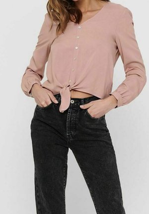 Žena v světle růžové košili s knoflíky přepásané v pase a vysokým pasem vystřihaných černých džínách s rukama v kapse.