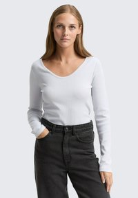 Lichtgrijze langarmige trui met ronde hals, geribbelde textuur, gecombineerd met hoge taille donkere jeans, model poseert met handen in zakken.