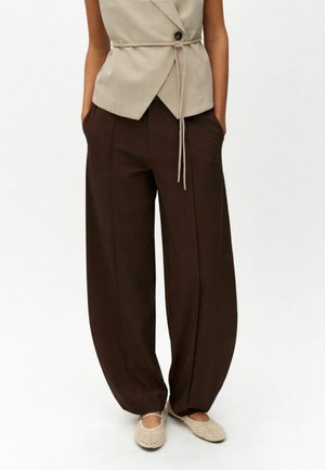 Beige ærmeløs slå-om top med bindebånd og mørkebrune bukser med brede ben, kombineret med flettede beige flade sko på en stående person.