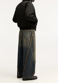 Overmatige donkere denim jeans met een rafelige, schuine naadontwerp; gecombineerd met een zwarte bomberjack met geribbelde kraag. Zwarte schoenen maken de look compleet.