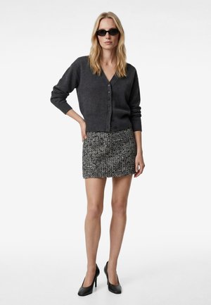 Kvinde iført sorte solbriller, mørkegrå cardigan med knapper, sort-hvidt mønstret mininederdel og sorte højhælede sko, stående med den ene hånd på hoften.