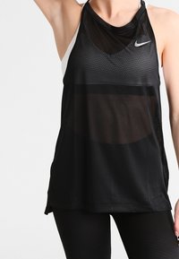 Mujer que lleva una camiseta sin mangas de malla negra de Nike sobre un sujetador deportivo blanco, combinada con leggings negros, de pie con un brazo levantado.