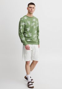 Grøn sweatshirt med hvide blomsterprint, kombineret med lysegrå shorts, hvide sokker og brune sandaler. Simpelt, afslappet design.