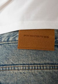 Patch en cuir marron avec l'inscription "PROHIBITED" embossée sur un jean en denim bleu clair porté sous une chemise blanche.