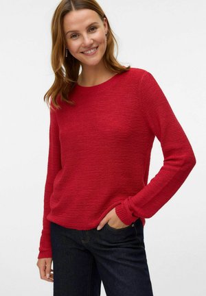 Pull en tricot rouge avec un col rond, des manches longues et une texture subtile. Porté sur un jean bleu foncé, avec une coupe décontractée.