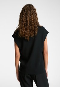 Top de punto negro de manga corta con un ajuste holgado y dobladillo acanalado; textura suave con costuras mínimas; vista trasera que muestra el cabello rizado y la simplicidad del diseño.
