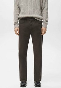 Hombre con pantalones rectos de color marrón oscuro, suéter gris claro de canalé y zapatos de cuero negros, de pie contra un fondo liso.