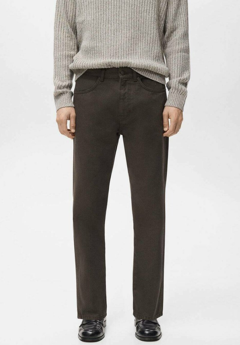 Hombre con pantalones rectos de color marrón oscuro, suéter gris claro de canalé y zapatos de cuero negros, de pie contra un fondo liso.