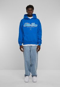 Sininen puuvillaneule, jossa on "Fake Love" -grafiikka vaaleansinisellä ja punaisella, kengurutasku, valkoisen T-paidan päällä, yhdistetty vaaleansinisiin farkkuihin.