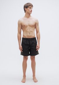 Μαύρα μαγιό σορτς με ελαστική μέση, κλείσιμο με κορδόνι και χρυσό γεωμετρικό σχέδιο στα πλαϊνά. Το μοντέλο στέκεται ξυπόλητο μπροστά σε απλό φόντο.