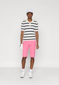 Polo de mangas curtas às riscas em azul-marinho e branco, combinado com calções cor-de-rosa. O modelo usa um boné branco e luva de golfe, com ténis brancos.
