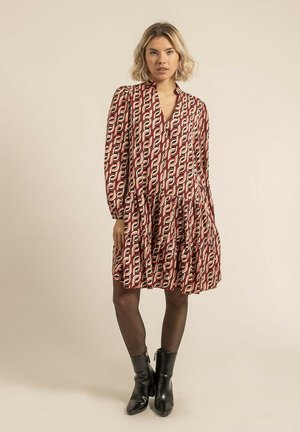 Robe à motifs rouge et beige avec des manches longues, un col en V et une jupe à étages. Portée avec des bottines noires, sur un fond neutre.