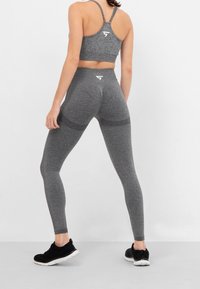 Szary stanik sportowy i legginsy, wykonane z teksturowanej tkaniny, z logo z tyłu. Czarne buty sportowe uzupełniają strój.