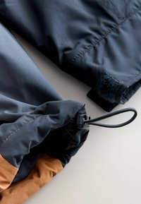 Material rezistent la intemperii de culoare neagră și maro, cu o deschidere elastică strânsă dotată cu un toggle și o bandă Velcro pentru închidere sigură.