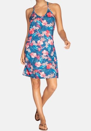 Vrouw in een blauw bloemenhemdjurk zonder mouwen met roze en witte bloemen, die zelfverzekerd in bruine sandalen loopt op een effen achtergrond.