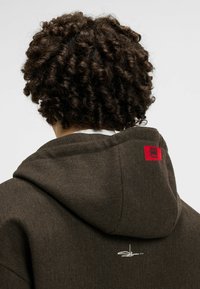 Brązowa bluza z kapturem w wzór jodełki z czerwoną metką logo na kapturze oraz białymi haftami w okolicy dekoltu. Teksturowana tkanina i luźny krój.