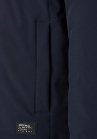 Nahaufnahme von einer dunkelblauen Jackentasche mit einem rechteckigen genähten Etikett, das auf der linken Unterseite "NORTHERN EXPD Since 1924" liest.