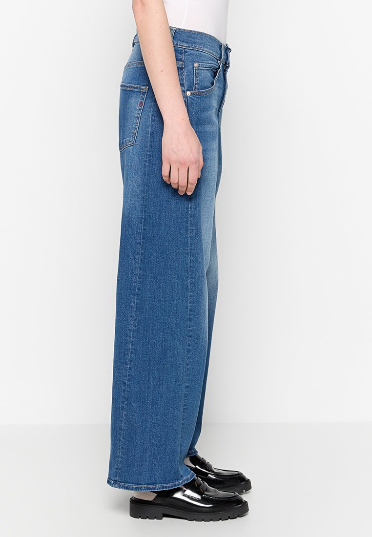 Weite blaue Denim-Jeans mit mittlerer Bundhöhe, ausgestattet mit fünf Taschen und dezenten Nähdetails. Kombiniert mit schwarzen Plateauschuhen.