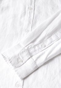 Camisa de lino blanca con cierre de botones en la parte delantera, mangas largas y botones de plástico transparente en los puños, con un sutil detalle de textura.