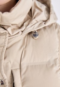 Bež puffer jakna s visokim ovratnikom, mekanom teksturom i detaljima od metalnih dugmadi. Uključuje zip džep sprijeda.