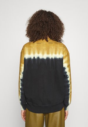 Persoon met krullend haar die een geel, zwart en wit tie-dye sweatshirt en olijfgroene broek draagt, van achteren gezien tegen een effen achtergrond.