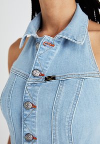 Robe en denim bleu clair avec un col, ornée de boutons argentés, d'accents de couture orange et d'une étiquette noire avec le nom de la marque.