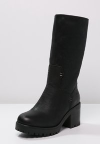 Bota negra hasta la rodilla con un eje de cuero suave, tacón grueso y suela de goma texturizada. Presenta acentos de costura mínimos.