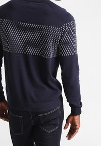 Granatowy sweter dzianinowy z teksturowaną, wzorzystą górną częścią, zdobioną małymi, białymi pionowymi liniami. Ściągacze u mankietów i na dole. Noszony z ciemnymi dżinsami.