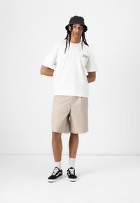 Witte katoenen t-shirt met tekst, beige shorts, zwarte bucket hat en zwarte sneakers met witte zolen en veters, staand op een effen achtergrond.