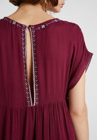 Bordó šaty s texturovanou tkaninou, zdobené květinovou výšivkou a detailem s otvorem na zádech. Rukávy ve tvaru motýla a sukně s nařasením.