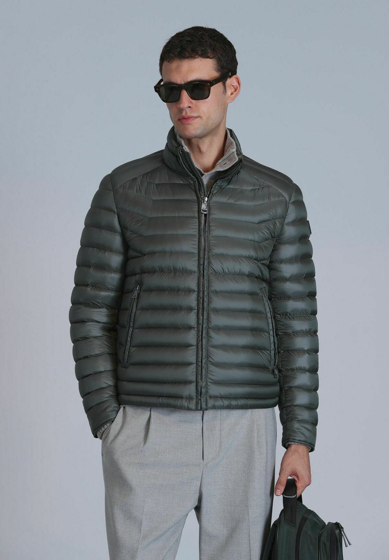 Doudoune grise avec texture matelassée, col haut et poches zippées. Portée avec un pantalon gris et tenant un sac vert foncé.