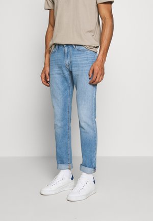 Mann trägt hellblaue, umgeschlagene Jeans, beige T-Shirt, weiße Socken und weiße Sneakers mit blauen Fersenakzenten, steht vor schlichtem Hintergrund.