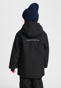 Barn som bär en svart vinterjacka med huva och en marinblå stickad mössa, sedd bakifrån mot en plain ljusbakgrund.