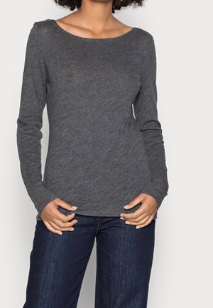 Personne portant une chemise à manches longues gris foncé et un jean bleu foncé, les mains posées près de la taille sur un fond uni.