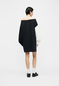 Γυναίκα με κοντά σκούρα μαλλιά που φοράει μαύρο φόρεμα έξωμο με ένα λευκό μανίκι, λευκές κάλτσες και μαύρα αθλητικά παπούτσια, στέκεται με την πλάτη προς τα εμπρός.