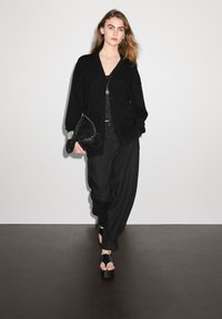 Svart cardigan med knappar, svart topp, lösa svarta byxor och platåsandaler. Modellen håller en texturerad svart väska och poserar mot en enkel bakgrund.