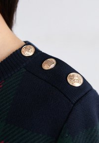 Marinblå tröja med ribbad krage, prydd med tre gyllene dekorationknappar på axeln och ett strukturerat mönster i mörkgrön.