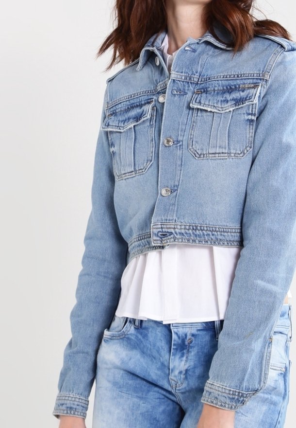 Giacca di jeans cropped in blu chiaro con due tasche sul petto, bottoni argentati e una texture leggermente vissuta. Indossata sopra una blouse bianca.