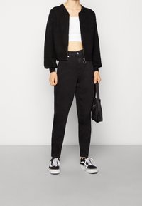 Czarna dzianinowa kurtka typu bomber, biała krótka bluzka na ramiączkach, czarne spodnie z wysokim stanem oraz czarne trampki. Modelka trzyma małą czarną torebkę.