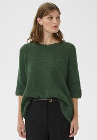 Grüner Strickpullover mit lockerem Sitz, dreiviertel Ärmeln und rundem Ausschnitt, strukturierte Oberfläche, kombiniert mit schwarzen, hoch taillierten Hosen.