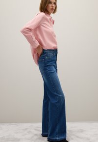 Donna con una camicia a righe rosse e bianche infilata in un paio di jeans alti blu, in piedi di lato contro un muro chiaro e semplice.